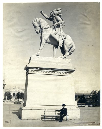 Vertikales, sepiafarbenes Foto einer Statue eines amerikanischen Indianers auf einem Pferd auf einem großen Sockel. Ein Mann im dunklen Anzug und Hut sitzt auf einer Bank am Sockel von Unknown